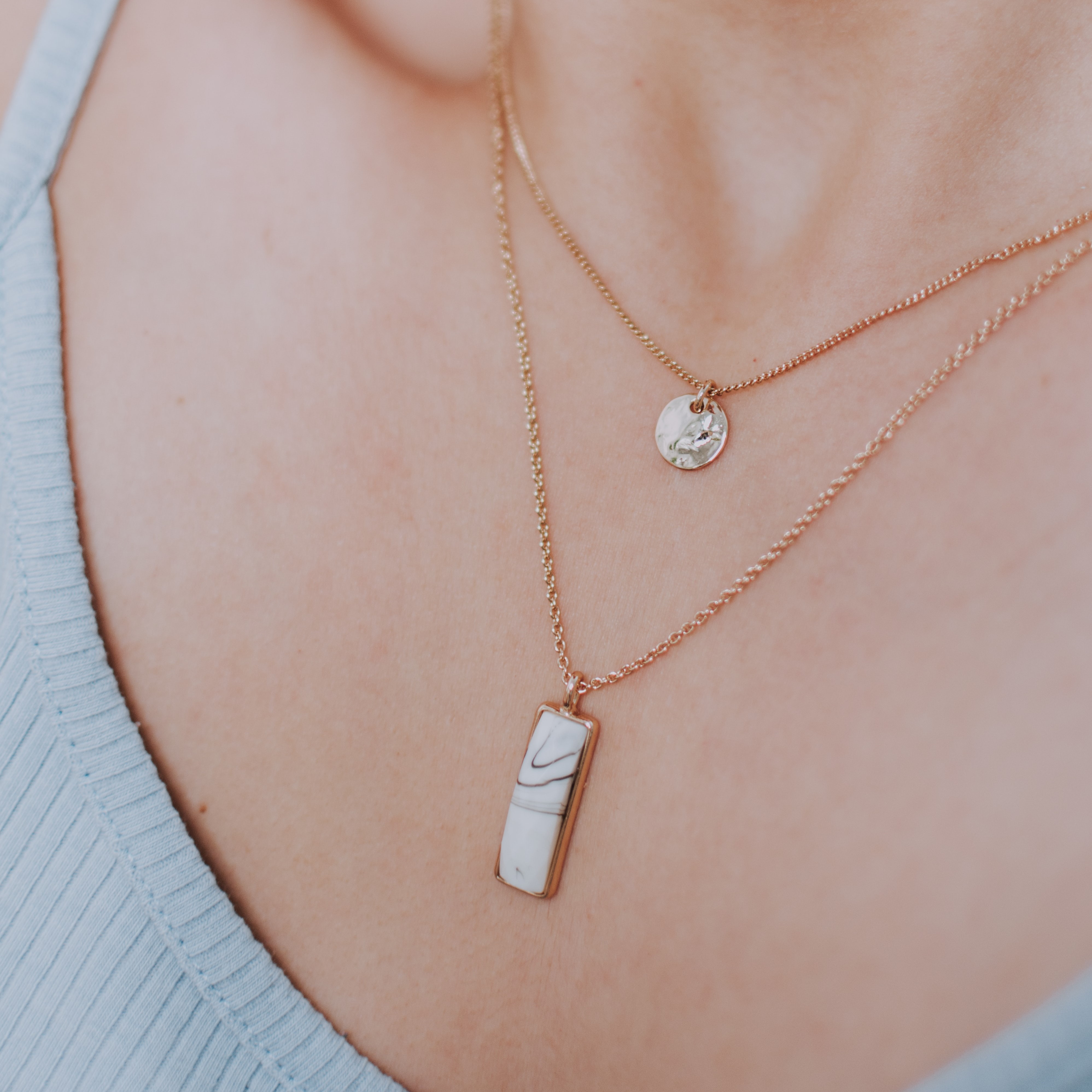 A thin gold necklace with two charms, one round and gold and second rectangular and with marbel texture